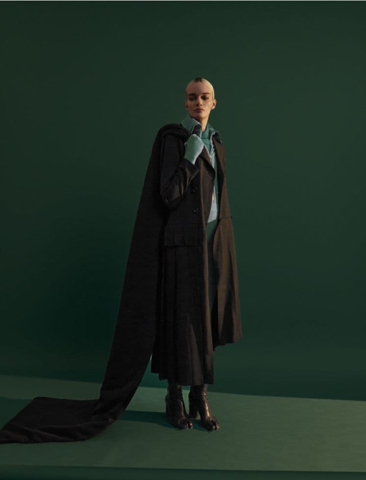 Model in a long dark coat with a trailing train and pale teal scarf, against a deep green backdrop.