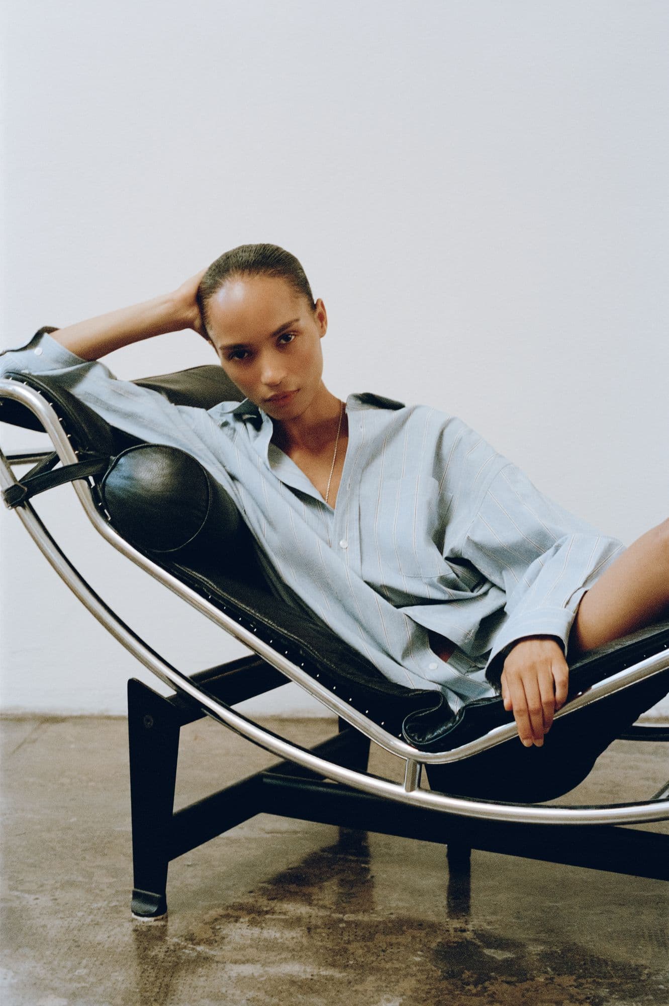 Model reclining on a Le Corbusier chaise longue in an oversized pale blue pinstripe shirt, against a bare white wall.
