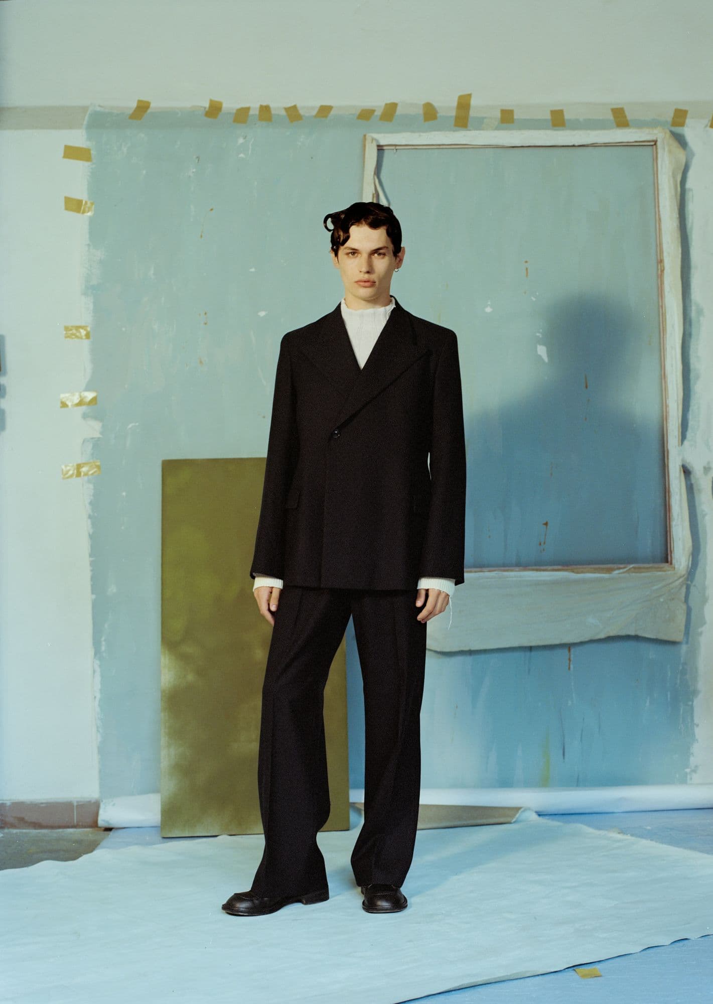 Model in a black tailored suit standing before a taped, paint-washed pale blue backdrop with a bare frame and gold panel.