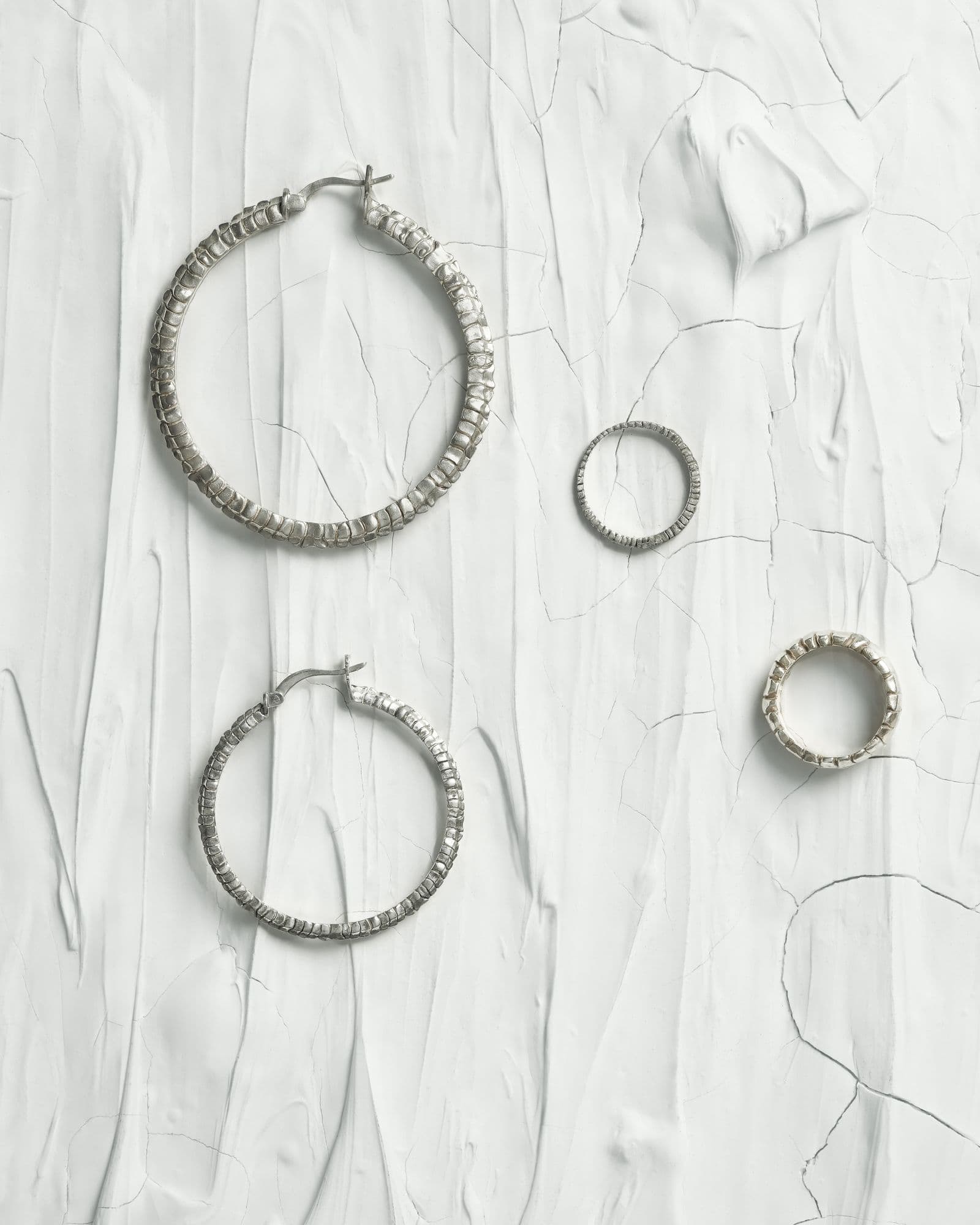 Textured silver hoop earrings and two rings laid out on creased white plaster, shot from above.